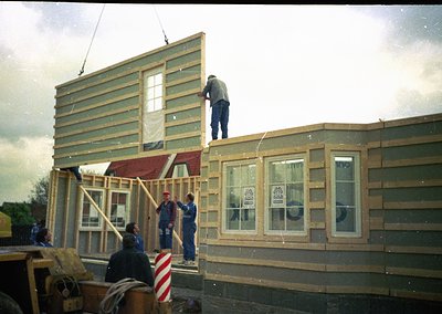 Two construction workers frame a two-story wooden house exterior, likely mid-1980s–1990s. Green siding and red roofing contra...