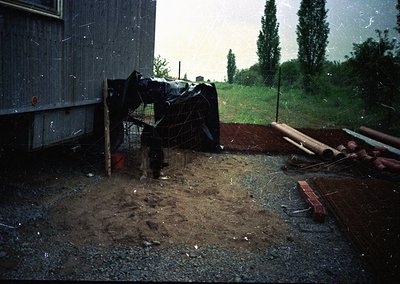 Vintage rural scene featuring a person in heavy rain gear standing near a makeshift fence and tarpaulin-covered structure. Mu...