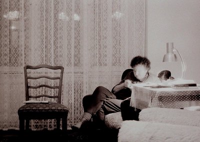 Mid-20th-century bedroom scene: child seated on patterned bedspread, engrossed in a book. Wooden chair with curved backrest a...