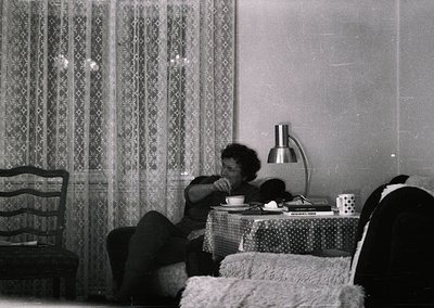 Mid-century bedroom scene featuring a person seated on a bed, drinking from a cup. Decor includes patterned curtains, a table...