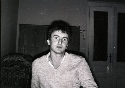 Blurred black-and-white portrait of a young man in a textured V-neck sweater, seated indoors near a wicker chair and open doo...