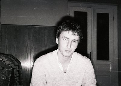 Portrait of a young man in a light sweater, captured in low-light indoor setting. Wooden door and patterned upholstery sugges...