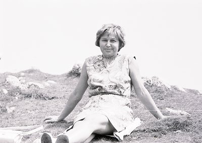 Portrait of a woman in a floral-patterned dress, seated outdoors on grassy terrain with rocky background. Mid-20th century fa...