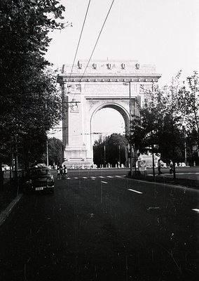 Neoclassical triumphal arch with intricate carvings and a central inscription, flanked by statues. Urban street scene with vi...