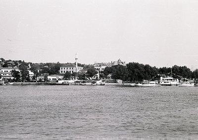 Black-and-white coastal scene featuring a prominent **minaret** and **waterfront buildings** along a shoreline. Small **ferri...