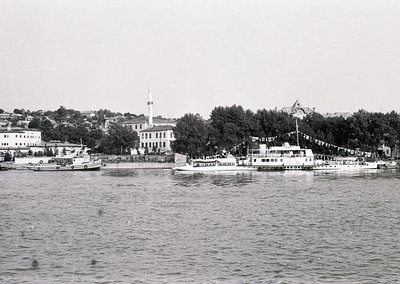 Black-and-white coastal scene featuring a waterfront promenade with a prominent building featuring a tall minaret and flagpol...