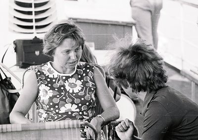 Black-and-white candid shot of two individuals engaged in conversation on a boat deck, likely mid-20th century. Woman in flor...