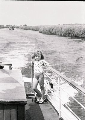 A young girl in a floral dress and sandals stands on a boat railing, smiling at the camera, with reeds and water in the backg...