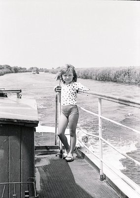 Mid-20th century girl in polka-dot blouse and shorts stands on a ship’s deck railing, gripping metal handrail. Waterway and d...