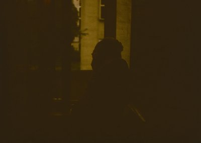 Silhouette of a person standing near a dimly lit, narrow alleyway with visible brick walls and a faintly lit doorway. Sepia-t...