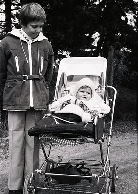 A 1970s-era child pushes a vintage stroller with a baby seated inside, both dressed in winter attire. The older child wears a...