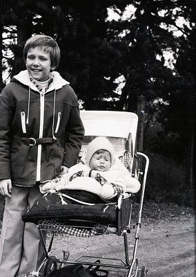 Vintage black-and-white photo of a young boy pushing a baby buggy with an infant dressed in a knitted outfit and hat. The boy...