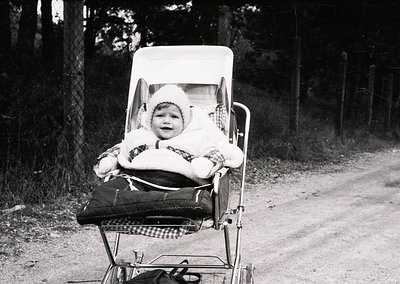 Vintage black-and-white photo of a child in a vintage baby carriage on a rural road, bundled in a knitted hat and fur-lined c...