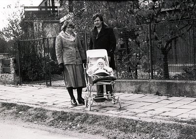Two women in 1960s-era attire pose outdoors beside a classic metal-framed baby stroller. The woman on the left wears a fur co...