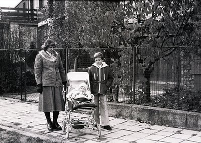 A mid-20th-century black-and-white snapshot captures a woman in a knee-length coat and scarf pushing a vintage pram, accompan...