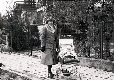 A woman in 1960s-era attire—coat, gloves, and hat—pushes a classic metal-framed pram with a sleeping infant. Residential stre...