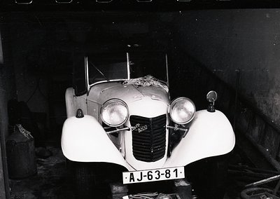 Classic 1930s-era roadster with streamlined bodywork, dual round headlights, and "AJ-63-81" license plate. Indoor setting sug...