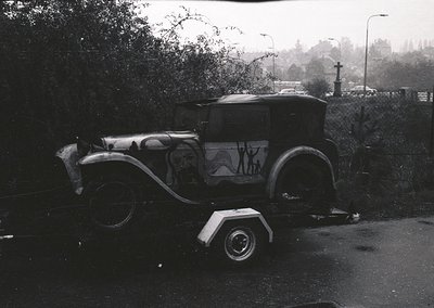 Vintage flatbed trailer towing a 1930s-era sedan with decorative mural depicting human figures, parked on a wet roadside. Sur...