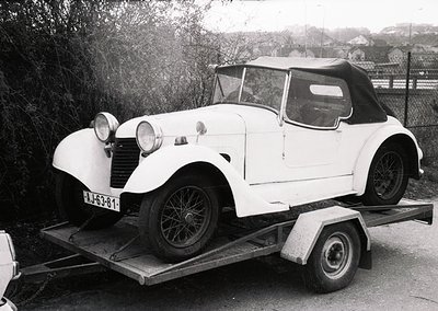 Classic white convertible roadster on trailer, likely a 1930s–1940s model, featuring rounded fenders, dual headlamps, and a v...
