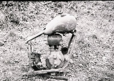 Vintage lawnmower engine with exposed cylinder and carburetor, set in overgrown grass. Likely mid-20th century agricultural e...