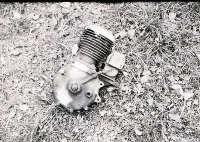 Vintage single-cylinder motorcycle engine in grassy outdoor setting, likely mid-20th century. Rust and wear suggest prolonged...