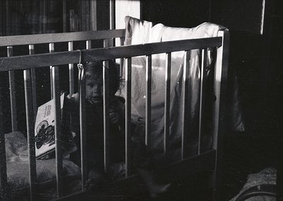 Child peering through wooden crib bars in dimly lit room, mid-20th century. Visible: vintage packaging (likely food) on left,...
