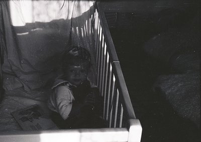 Black-and-white candid of a child peering through a crib’s vertical bars, clutching a booklet with a visible illustration. So...