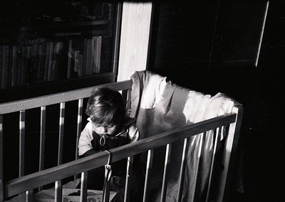 A young child sits in a wooden crib, leaning forward with hands on the bars, gazing intently. Bookshelves filled with books l...