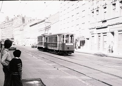 Vintage streetcar () navigating urban tracks in mid-20th century cityscape. Pedestrians observe from sidewalk beside mid-rise...