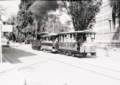 Vintage steam-powered tram with numbered cars (10 & 6) traveling on urban tracks, emitting visible smoke. Passengers stand on...