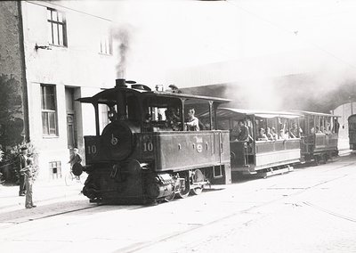 Vintage steam locomotive pulling open-air passenger cars, likely a tourist train, in early 20th-century Europe. Smoke billows...
