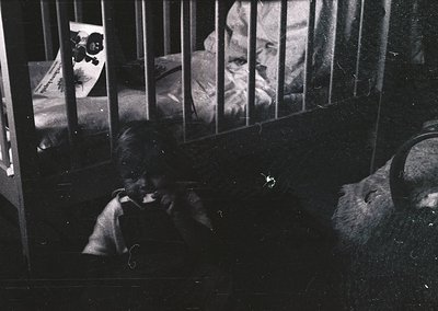 Black-and-white prison cell interior with barred window revealing two inmates. One sits on a bunk, reading a newspaper; the o...