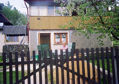 Two children pose in front of a mid-20th-century residential house with a yellow upper floor and grey lower walls. Green plas...