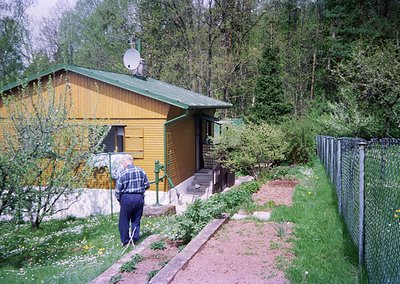 A man in a checkered shirt and cap tends to a small, rustic wooden cabin with a green roof, set in a forested area. Satellite...
