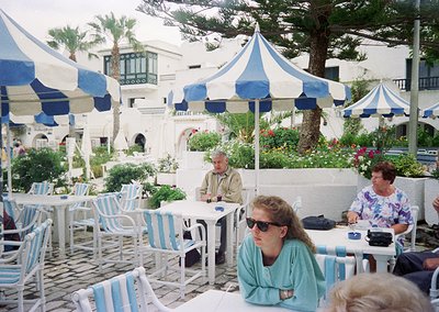 Mid-1990s seaside café with Mediterranean architecture—whitewashed walls, arched doorways, and lush greenery. Patrons dine un...