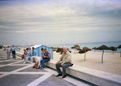 Vintage seaside promenade with mid-century concrete seating. Elderly man in light jacket sits on a curved concrete ledge, ove...