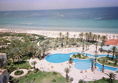 Aerial view of a mid-20th-century seaside resort with curved infinity pool, palm-dotted lawns, and golden beach stretching to...