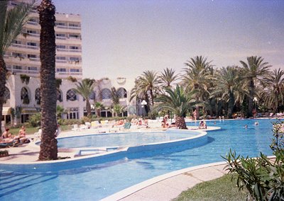 Mid-20th century seaside resort pool with palm-draped perimeter, sunbathers, and multi-story hotel in background. Architectur...