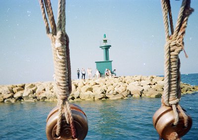 Vintage seaside lighthouse perched on a breakwater of stacked rocks, framed by rusted metal mooring rings. Small group of peo...