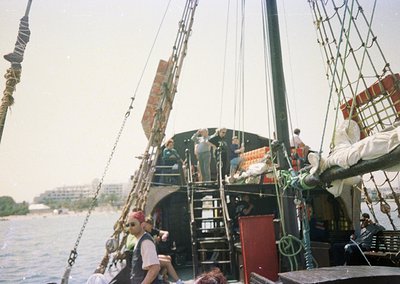 Vintage wooden sailing ship with three masts, likely a replica of a 17th-century East Indiaman, docked near an urban waterfro...