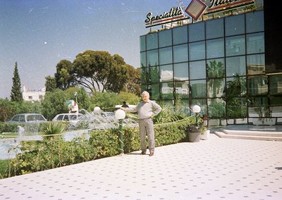 Man in light-colored suit waters lush courtyard garden beside modernist building with "Specialità" signage, likely a 1970s It...
