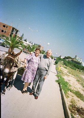 Couple posing beside a donkey-drawn cart in a coastal urban setting, likely Mediterranean. Woman in patterned dress, man in l...