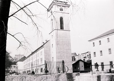 Mid-20th century urban scene featuring a tall, square clock tower with a bell and decorative cornice, likely part of a public...