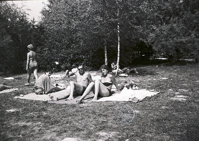 Mid-20th century beach scene: five individuals lounging on towels in a wooded area, likely 1950s–1960s. Minimalist swimwear a...