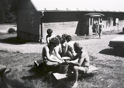Mid-20th century group photo: five individuals in swimwear playing with a bucket near a rustic wooden cabin. Classic 1950s-60...