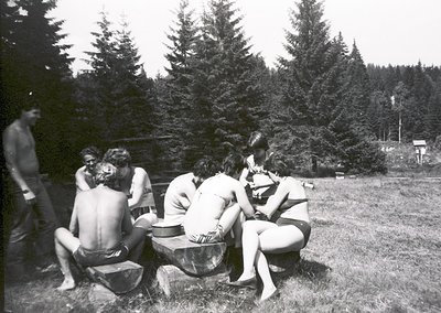 Group of six people in 1960s-70s outdoor setting, seated around a large metal drum. Four women in swimsuits, two men shirtles...