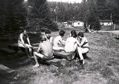 Six individuals in swimwear pose casually on a rustic wooden bench beside a forested lake, mid-20th century. Lush greenery an...