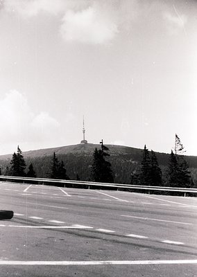 Mid-century roadside view of a tower atop a forested hill, likely a telecommunications or observation structure. Asphalt road...