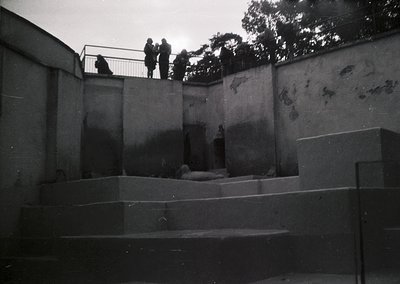 Minimalist concrete courtyard with geometric walls and steps, featuring a lone reclining figure. Three silhouetted figures st...