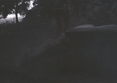 Vintage black-and-white photo of a dimly lit interior with faint silhouettes. A single figure stands near a window, backlit b...